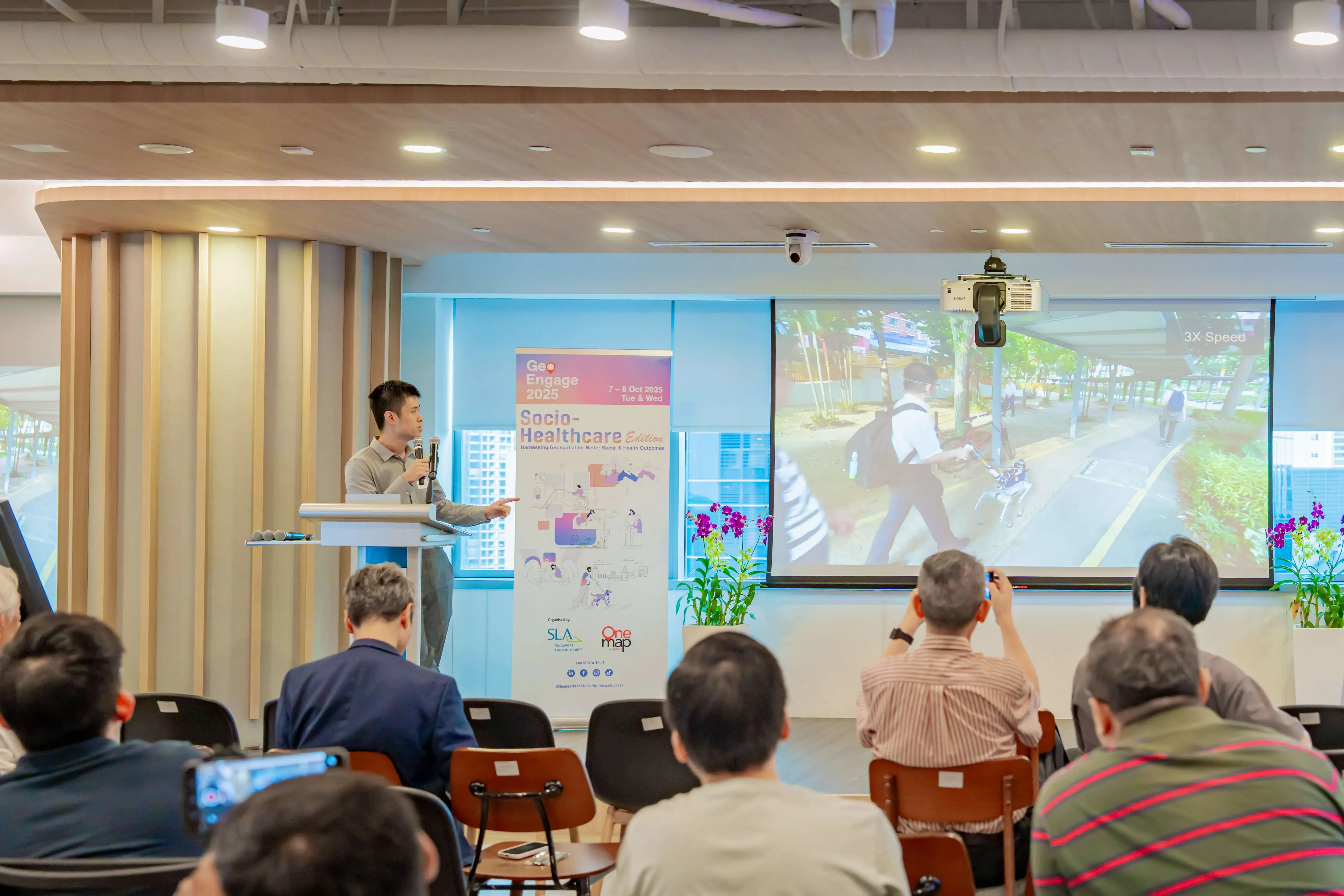A man speaks at a podium at the GeoEngage 2025 Socio-Healthcare event while attendees watch a projected street video.